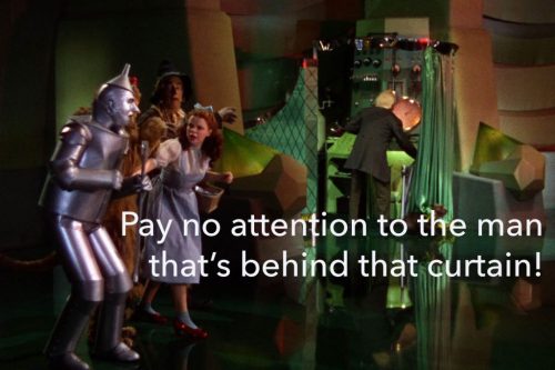 Photo shows scene from Wizard of Oz, with Dorothy, Tin Man, Lion, Scarecrow, as they pull back the screen to see the Wizard of Oz, who is a small man using a large microphone and lights to look "great and powerful"
